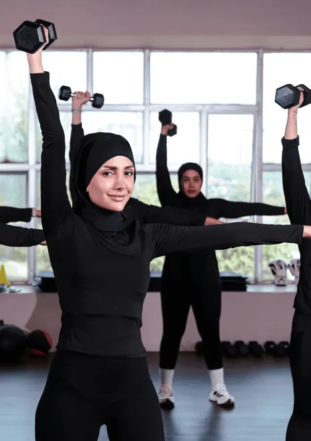 A class of women in hijab and black activewear press dumbbells overhead in a bright studio.