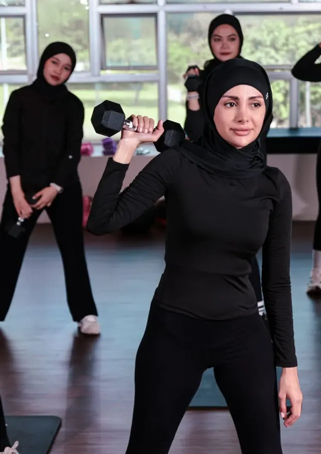 A woman in a black hijab lifts a pair of dumbbells while a class trains behind her.