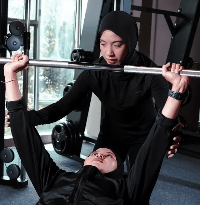 A coach in a black hijab stands over a member on a bench, spotting her barbell press.